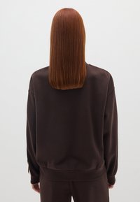 Sudadera marrón extragrande hecha de una suave mezcla de algodón, con cuello redondo y puños de canalé. Vista desde la parte posterior con el cabello lacio.