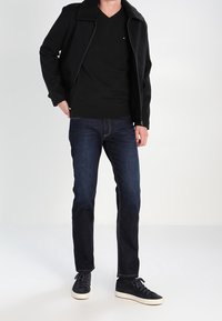 Svart zip-up jacka med fårskinnskrage, matchad med en svart V-ringad tröja, mörkblåjeans och svarta sneakers med vita sulor.