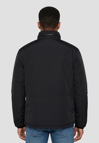 Giacca imbottita nera con collo alto, caratterizzata da una texture liscia e un design minimalista. Vista da dietro con jeans blu.