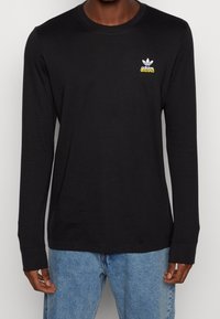 Svart långärmad t-shirt i bomull med rund halsringning. Har en vit Adidas-logotyp och gul text på vänster bröst. Avslappnad passform.