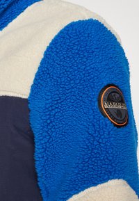 Fleecejakke med blått og kremfarget stoff, teksturert finish, og et svart og oransje logo-patch på overarmen.