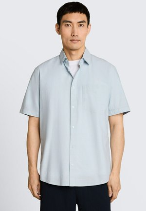 Homme portant une chemise bleu clair à manches courtes boutonnée sur un T-shirt blanc, debout devant un fond clair uni.