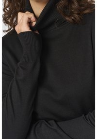 Personne aux cheveux bouclés portant un pull à col roulé côtelé noir, tenant le col avec une main près du menton.
