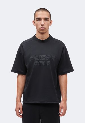 Jeune homme aux cheveux courts portant un t-shirt noir Adidas Golf, debout face à un fond clair uni.