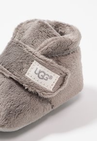 Grijze peuterboot gemaakt van zacht pluche materiaal, met een klittenbandsluiting en een UGG-logo patch. Rond vorm met een comfortabele kraag.