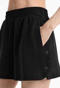 Zwarte shorts met een elastische tailleband, zijzakken en decoratieve knopenaccenten aan de zoom. De stof lijkt lichtgewicht en glad te zijn.