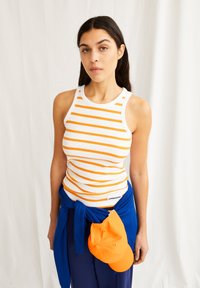 Weißes Tanktop mit orangefarbenen horizontalen Streifen, kombiniert mit einem blauen Stoff, der um die Taille gebunden ist, und einer orangefarbenen Kappe, die darauf ruht.