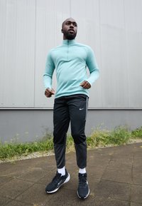 Jasnoniebieska, długa bluzka sportowa z zamkiem, w zestawie z czarnymi spodniami Nike i czarnymi sneakersami. Poza do biegania z fakturowanym materiałem.