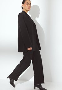 Črn prilagojen blazer in hlače z ohlapnim krojem, ostrimi reverji in kontrastnimi teksturami, v kombinaciji z visokimi črnimi škornji.