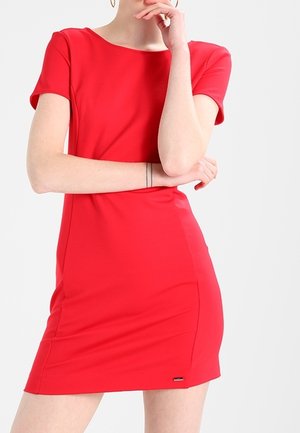 Kobieta w dopasowanej czerwonej sukience z krótkim rękawem, stojąca z jedną ręką skrzyżowaną, a drugą przy brodzie, na jednolitym tle.