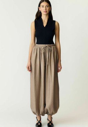 Femme portant un haut noir sans manches et un pantalon ballon beige ample avec des liens aux chevilles, debout devant un fond blanc uni.
