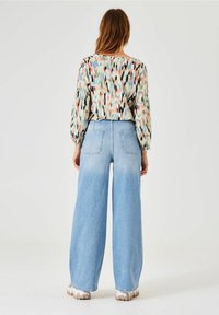 Blusa con estampado floral y mangas abullonadas, combinada con jeans de tiro alto y pierna amplia de color azul claro, que cuentan con bolsillos traseros y dobladillo deshilachado.