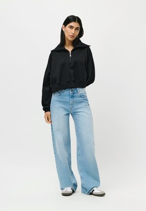 Kvinne i svart halv-zip genser, lyseblå vide jeans, hvite sneakers, som står mot en enkel lys bakgrunn.