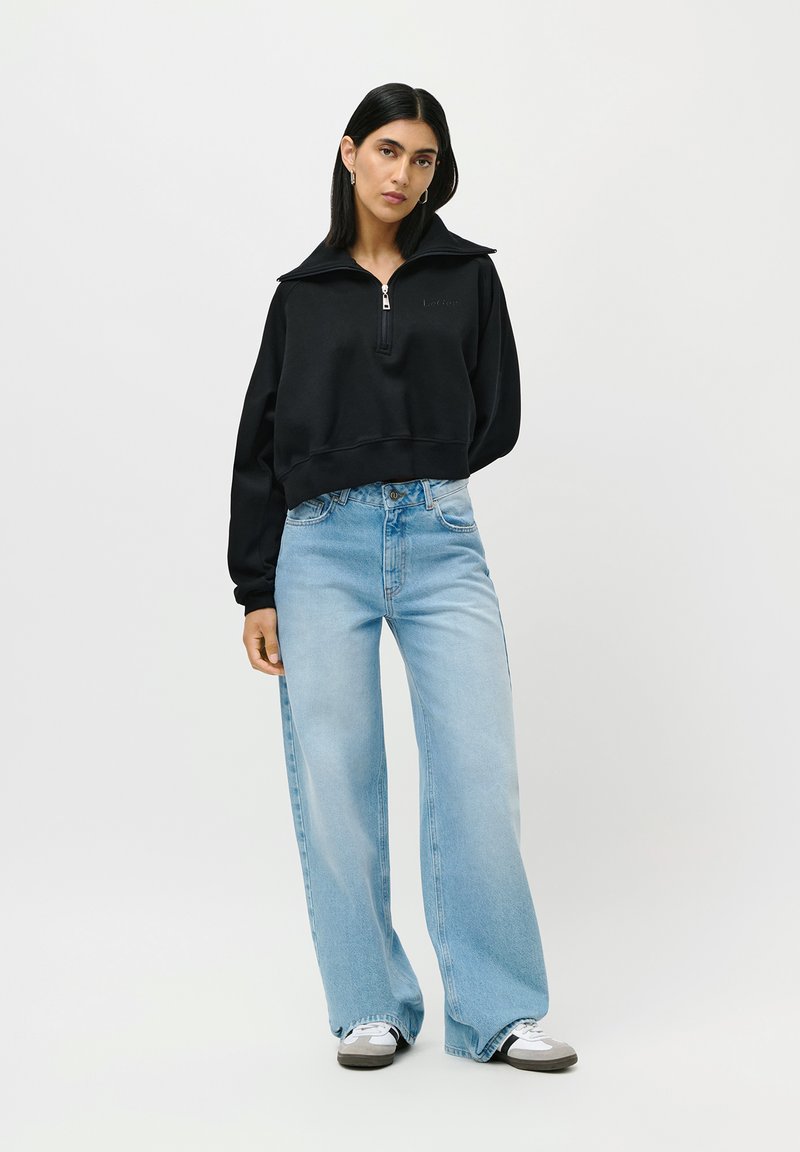 Mujer vestida con sudadera negra de media cremallera, jeans de pierna ancha de color azul claro, zapatillas blancas, de pie contra un fondo claro y liso.