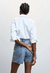 Camisa blanca de manga larga con los puños doblados, combinada con pantalones cortos de mezclilla de lavado claro. Tejido texturizado, diseño ajustado y bolsillos traseros visibles.