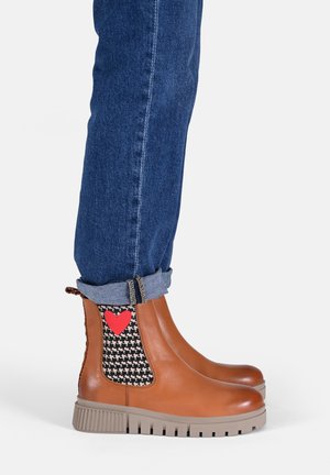 Bottines marron avec un panneau latéral à motif pied-de-poule noir et un détail en forme de cœur rouge. Semelle en caoutchouc avec des rainures, associées à un jean à revers.
