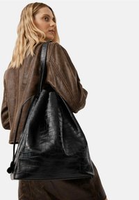 Donna con capelli biondi che indossa una giacca di pelle marrone e porta sulla spalla una grande borsa tote nera in pelle con motivo coccodrillo.