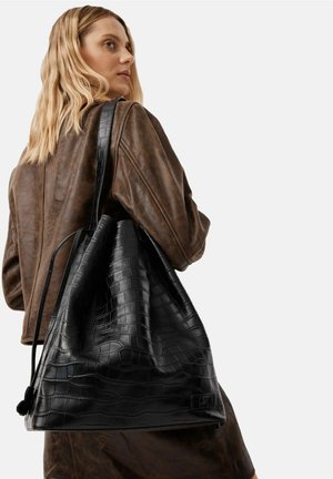 Frau mit blonden Haaren, die eine braune Lederjacke trägt und eine große, schwarze, krokodilgeprägte Ledertasche über der Schulter trägt.