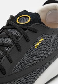 Zapatilla negra y gris con parte superior de malla, acento amarillo redondo, cordones planos y suela de goma texturizada; presenta la marca 'GEOX' en el lateral.