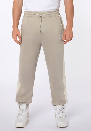 Beige Sweatpants mit weißen Seitenstreifen, elastischem Bund mit Kordelzug, vorderen Taschen, Bündchenabschlüssen und weißen High-Top-Sneakern.