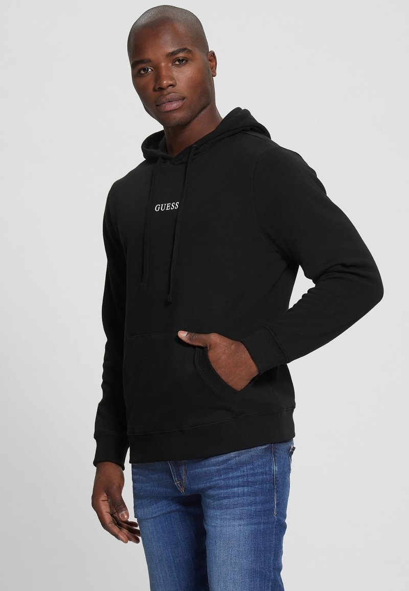 Black hoodie with a front pocket and drawstring hood. "GUESS" logo in white on the chest. Paired with blue jeans. Smooth texture.