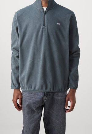 Mann trägt einen dunkelgrauen Fleecepullover mit Viertelreißverschluss und ausgewaschene schwarze Jeans, steht vor einem neutralen Hintergrund.