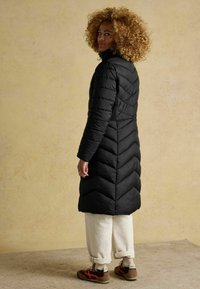 Melna pūkainā jaka ar chevron šuvēm, augstu apkakli un garām piedurknēm, kopā ar baltām biksēm ar uzsistām piedurknēm un brūniem sporta apaviem ar sarkaniem akcentiem.