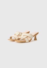Cream-colored heeled sandals with cross-straps in textured fabric, tan accents, open toe design, and a slender, tapered heel.
