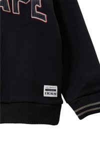 Felpa nera con scritte ricamate. Presenta un orlo e polsini a costine, e un'etichetta bianca con testo nero sul lato sinistro in basso.