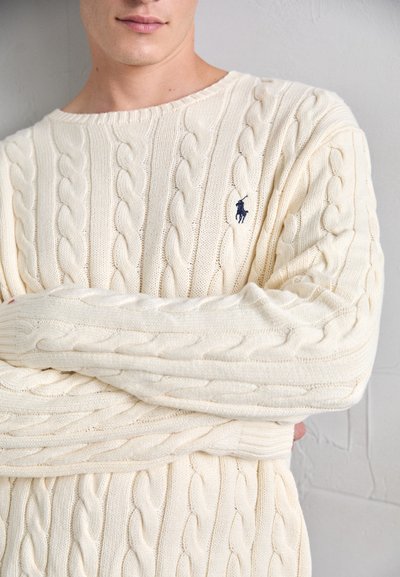 Man wearing a cream cable-knit sweater with a navy embroidered horse logo, standing with arms crossed against a textured white wall.