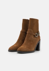 Botine din suede maro, până la gleznă, cu vârf conic, partea superioară dintr-un material similar cu o ciorap, și detalii decorative din metal pe curea. Tocuri pătrate adaugă stabilitate.