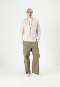 Ljusgrå zip-up hoodie, vit t-shirt, olivgröna vida byxor och vita sneakers. Kollen är beige och tyget ser mjukt och avslappnat ut.