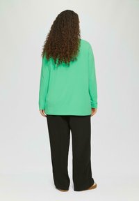 Groene blouse met lange mouwen en een ontspannen pasvorm, gecombineerd met zwarte wijd uitlopende broek. De blouse heeft een gladde textuur en een rechte zoom.