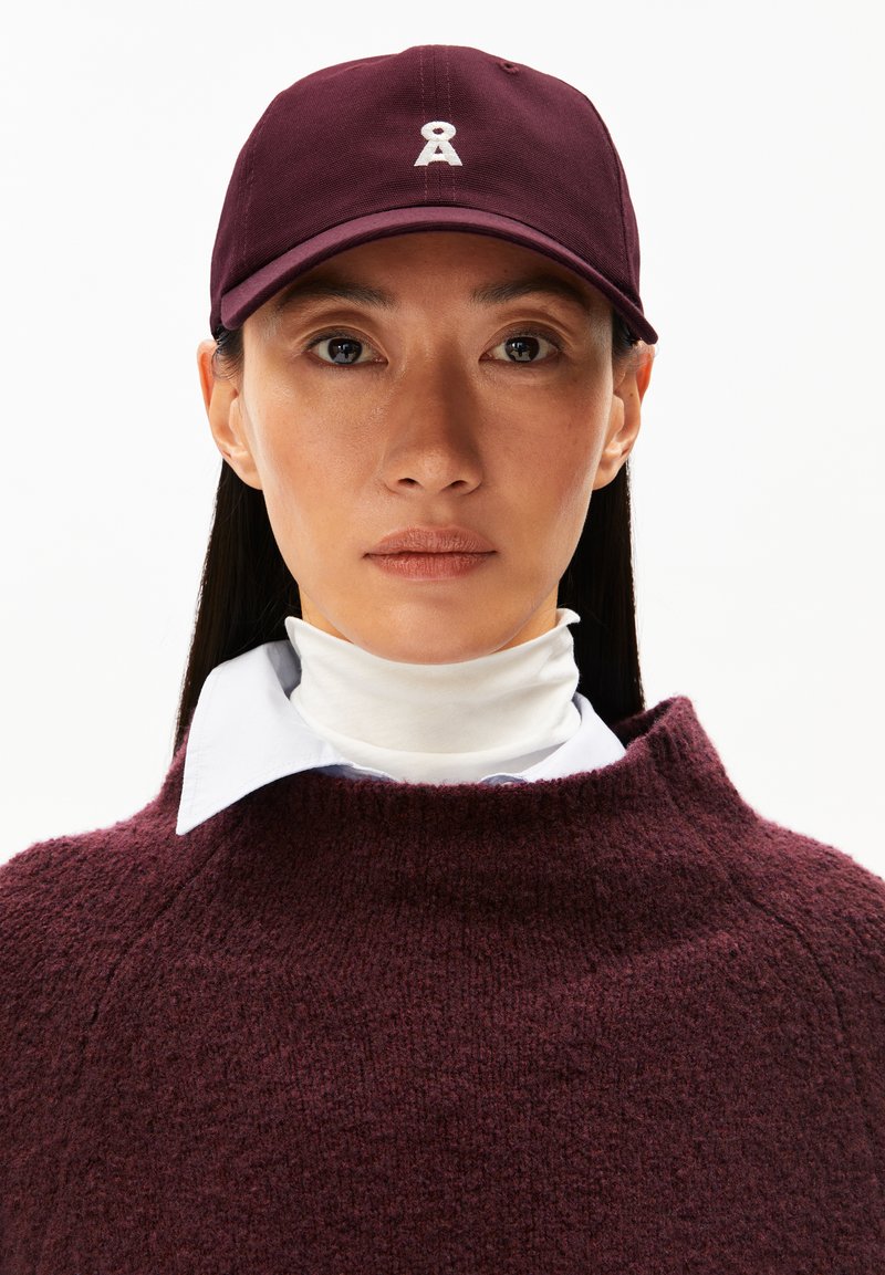 Bordeauxfarbige Mütze mit gebogenem Schirm und gesticktem Logo, kombiniert mit einem Rollkragenhemd. Der Pullover hat ein strukturiertes Strickdesign.