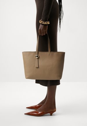 Borsa tote in pelle beige con due manici, caratterizzata da un elemento decorativo rotondo in oro. Il modello indossa un abito marrone a coste e décolleté marroni a punta.