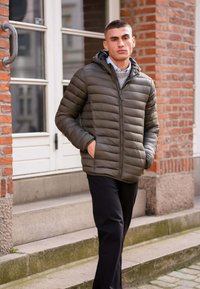 Polstret olivengrøn jakke med lynlås foran, hætte og vandret quiltet design, båret over en grå sweater og sorte bukser. Murstensvæg i baggrunden.