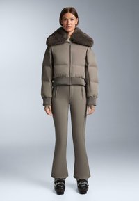 Párnázott olívazöld dzseki műszőrme gallérral, cipzáras záródással és bordázott mandzsettákkal, hozzá illeszkedő olívazöld nadrággal és fekete csizmákkal párosítva.