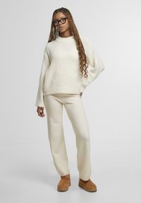 Krémový svetr s texturou a dlouhými rukávy, v kombinaci s krémovými kalhotami a hnědými semišovými platformovými botami. Modelka s copy stojí na bílém pozadí.