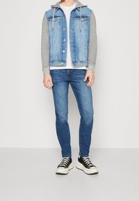 Jeansväst med grå hoodieärmar, framfickor, mörkblå jeans och svarta sneakers med vit sula. Avslappnad outfit med lager av texturer.