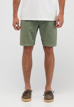 Mann trägt olivgrüne Shorts, weißes T-Shirt und braune Freizeitschuhe, steht vor einem schlichten hellen Hintergrund.