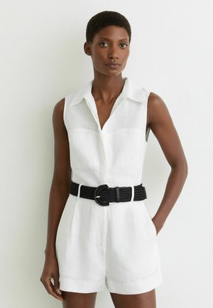 Femeie îmbrăcată într-un combinezon alb fără mâneci cu guler și o curea neagră împletită, stând împotriva unui perete alb simplu, cu o mână în buzunar.