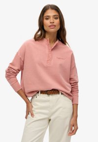 Top henley de manga larga rosa con tres botones y logotipo bordado, combinado con pantalones de color crema y un cinturón trenzado marrón.
