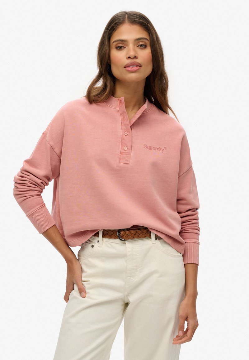 Top henley de manga larga rosa con tres botones y logotipo bordado, combinado con pantalones de color crema y un cinturón trenzado marrón.