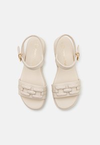 Sandales beiges avec une tige en cuir lisse, présentant un design de bride géométrique épaisse et une boucle de cheville réglable. Logo doré sur la semelle intérieure.