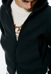 Felpa nera con zip realizzata in tessuto morbido, dotata di cappuccio, tasche frontali e logo ricamato. Indossata sopra un maglione crema con un motivo marrone.