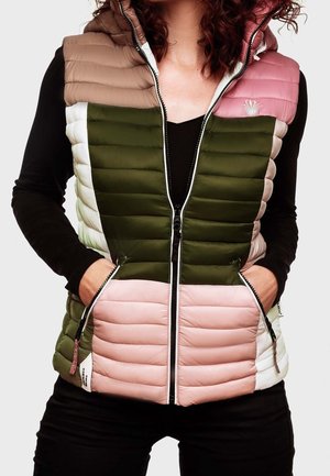 Vrouw draagt een mouwloos, meerkleurig gewatteerd vest met olijfgroene, roze, beige en witte blokken en een zwarte longsleeve eronder.