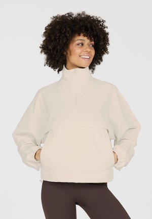 Kvinde med krøllet hår smilende, iført en lys beige pulloverjakke med høj krave og mørke leggings, hænderne i lommerne.
