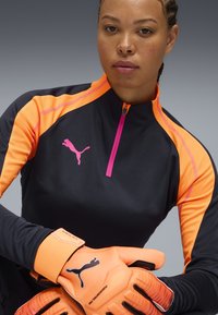 Maglietta sportiva nera con vivaci accenti arancioni e una zip rosa; presenta il logo Puma. Guanti arancioni vivaci con dettagli testurizzati.
