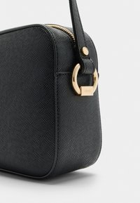 Černá texturovaná kabelka s zaobleným tvarem, zlatými kovovými detaily a zipovým uzávěrem. Obsahuje značkovou visačku a nastavitelný popruh.