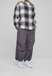 Osoba w oversize'owych szarych spodniach cargo, białych trampkach i zapinanej na zamek czarno-biało-szarej kurtce w kratę, stojąca z rękami w kieszeniach.